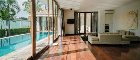 A spacious living room with a large window overlooking a swimming pool, a flat-screen TV mounted on the wall, and a beige sofa.