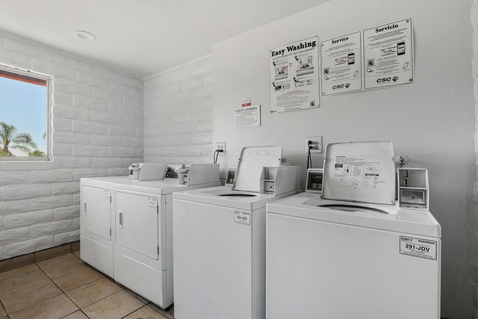A laundry area with two washing machines and a dryer, a window with a view of palm trees, and informational posters on the wall.