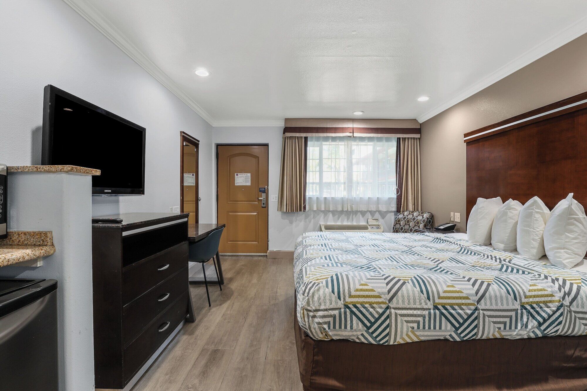 Habitación de hotel con cama, escritorio con televisor de pantalla plana, silla, puerta de madera y ventana con cortinas.