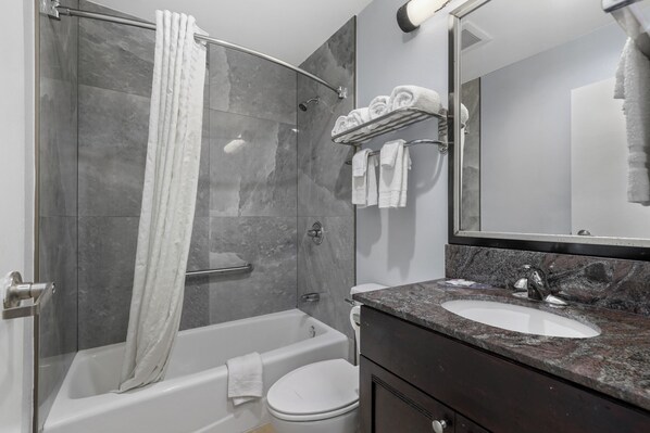 A modern bathroom with a glass-enclosed shower, a white bathtub, a toilet, and a vanity with a granite countertop.