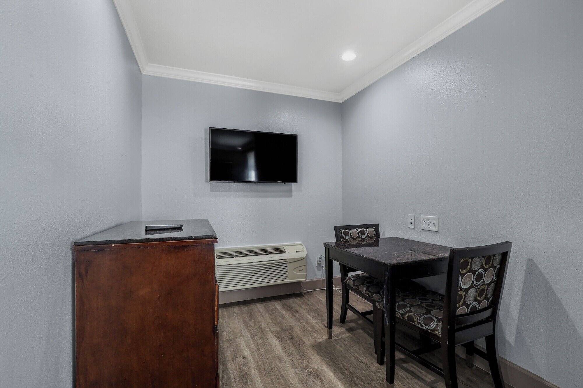 A small room with a flat-screen TV mounted on the wall, a wooden desk, a table with chairs, and a built-in air conditioning unit.