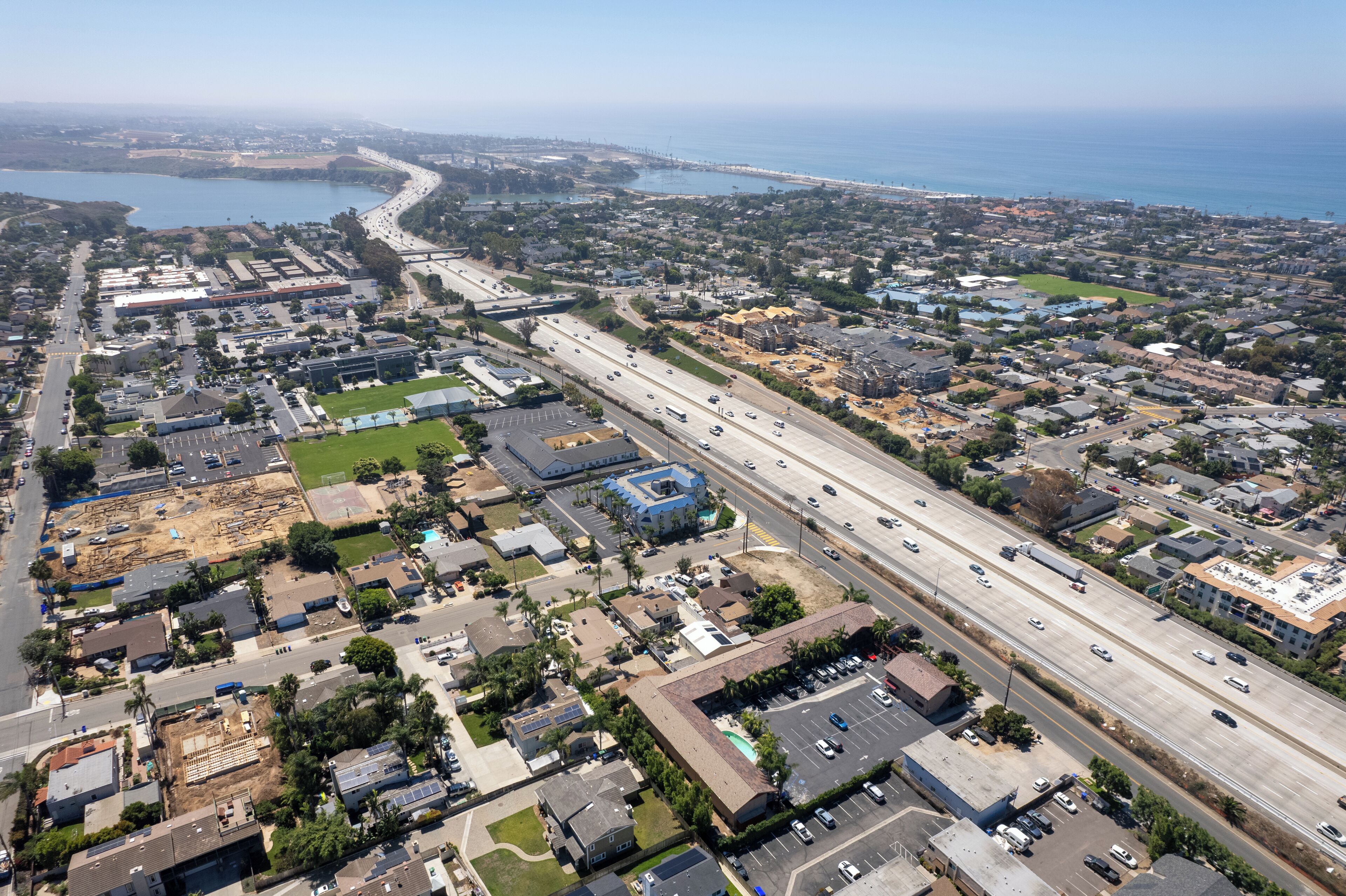An aerial view of a coastal urban area with a major highway, residential buildings, and a large body of water.