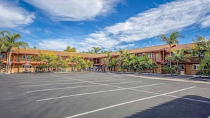 A parking lot with a building featuring a red-tiled roof and multiple balconies, surrounded by palm trees.