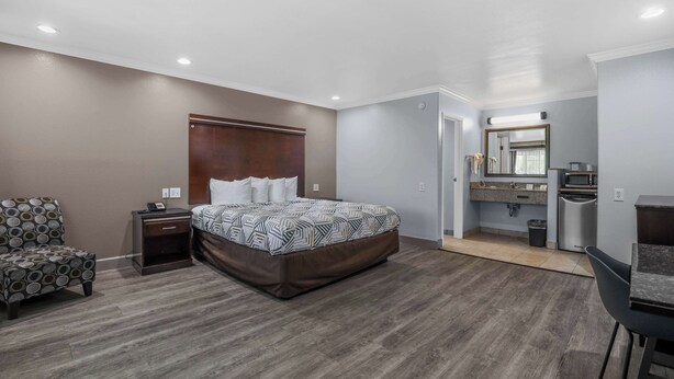 A bedroom with a bed, nightstand, chair, and a kitchenette area with a microwave and sink.