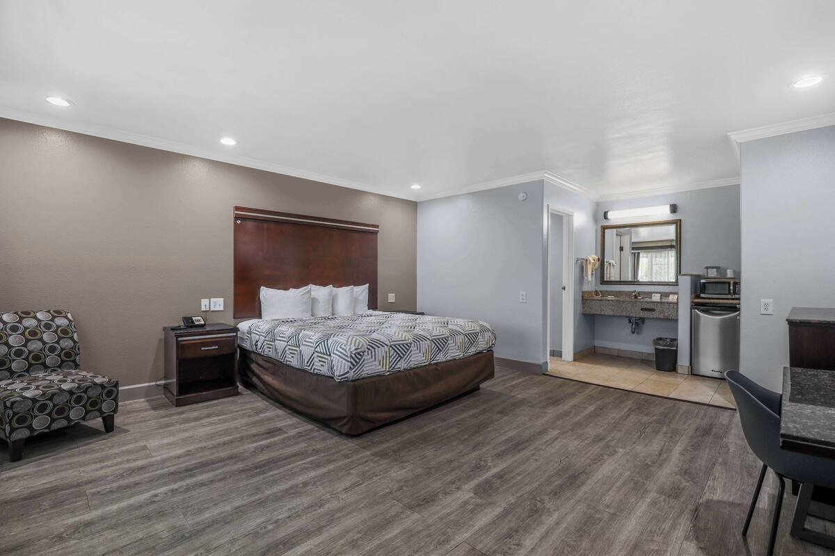 A bedroom with a bed, nightstand, chair, and a kitchenette area with a microwave and sink.
