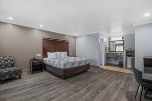 A bedroom with a bed, nightstand, chair, and a kitchenette area with a microwave and sink.