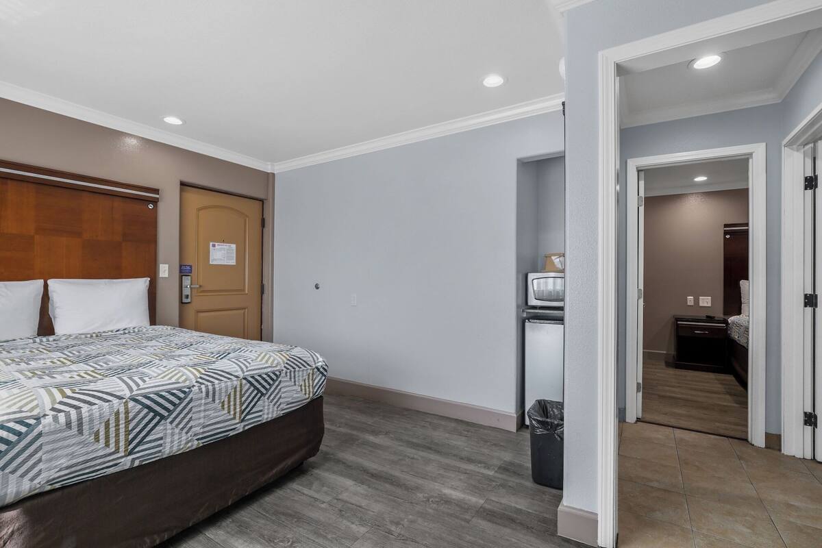 A bedroom with a bed, a wooden headboard, a door, a small kitchenette, and a trash bin.
