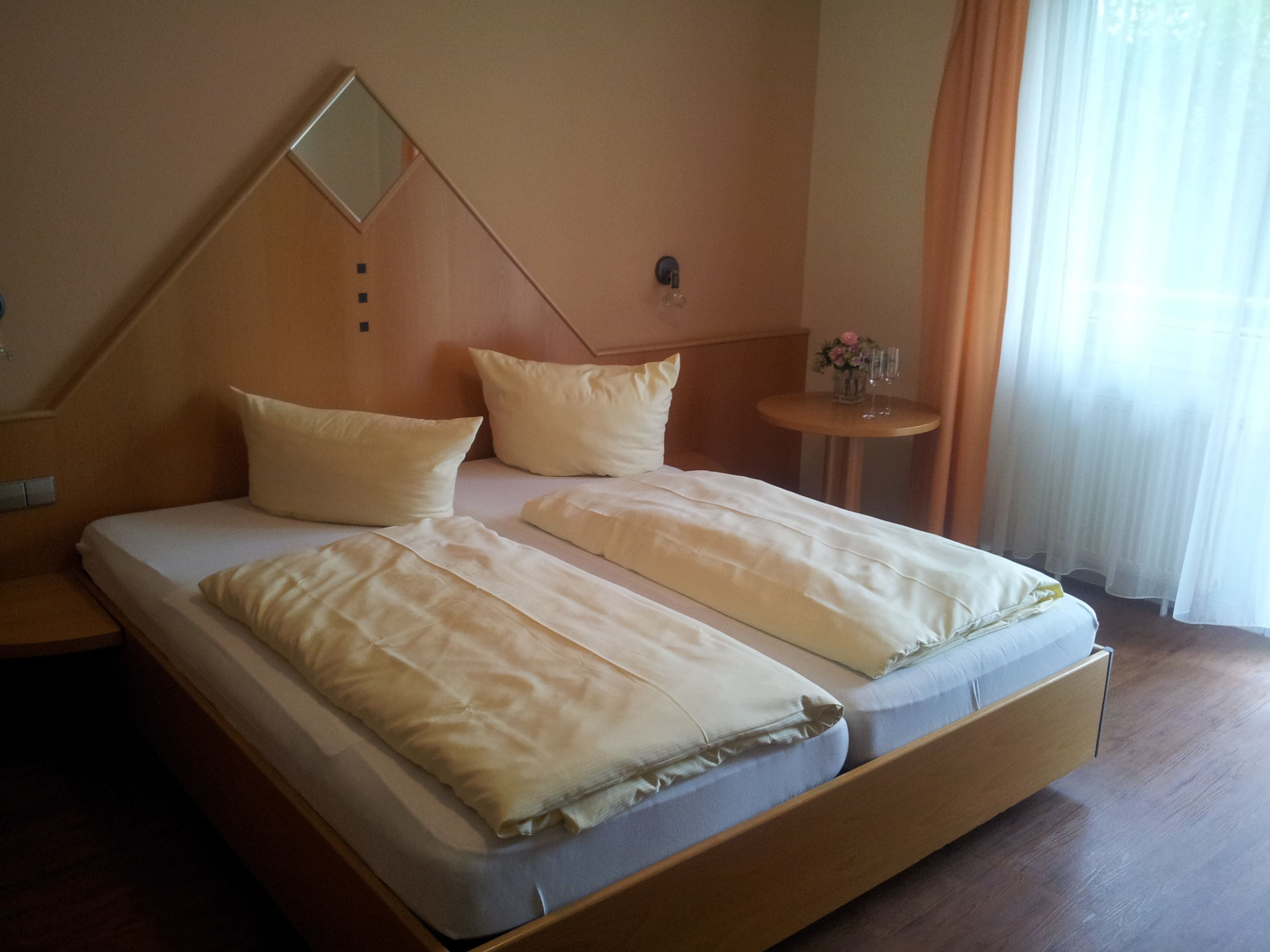 A double bed with a wooden frame, two pillows, a bedside table with a vase of flowers, and a window with curtains.