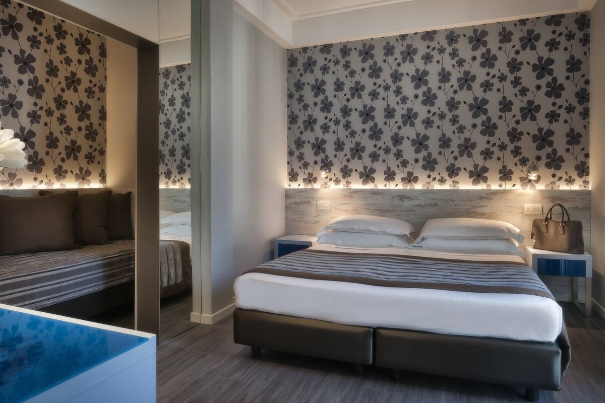 A modern hotel room with a large bed, bedside tables, a mirror, and floral wallpaper.