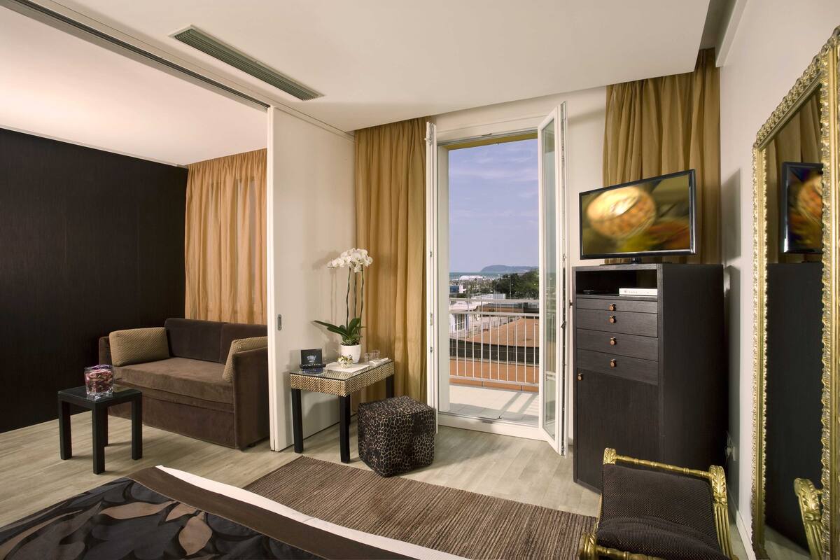 A hotel room with a sofa, a small table with a vase of flowers, a television on a stand, a wardrobe, and a view of the outdoors.