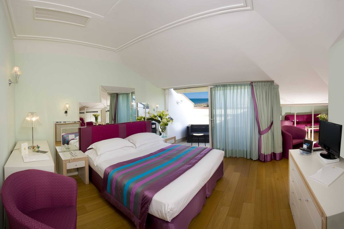 A hotel room with a large bed, a desk with a TV, a chair, and a balcony with a view of the sea.