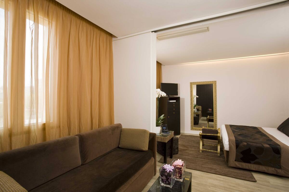 A hotel room with a sofa, a bed, a small table with decorative items, a television, and a mirror.