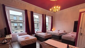A hotel room with three beds, a wooden desk, a chandelier, and a view of buildings through the windows.
