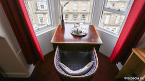 A corner room with a wooden table, a chair with a patterned cushion, and a vase with dried plants.