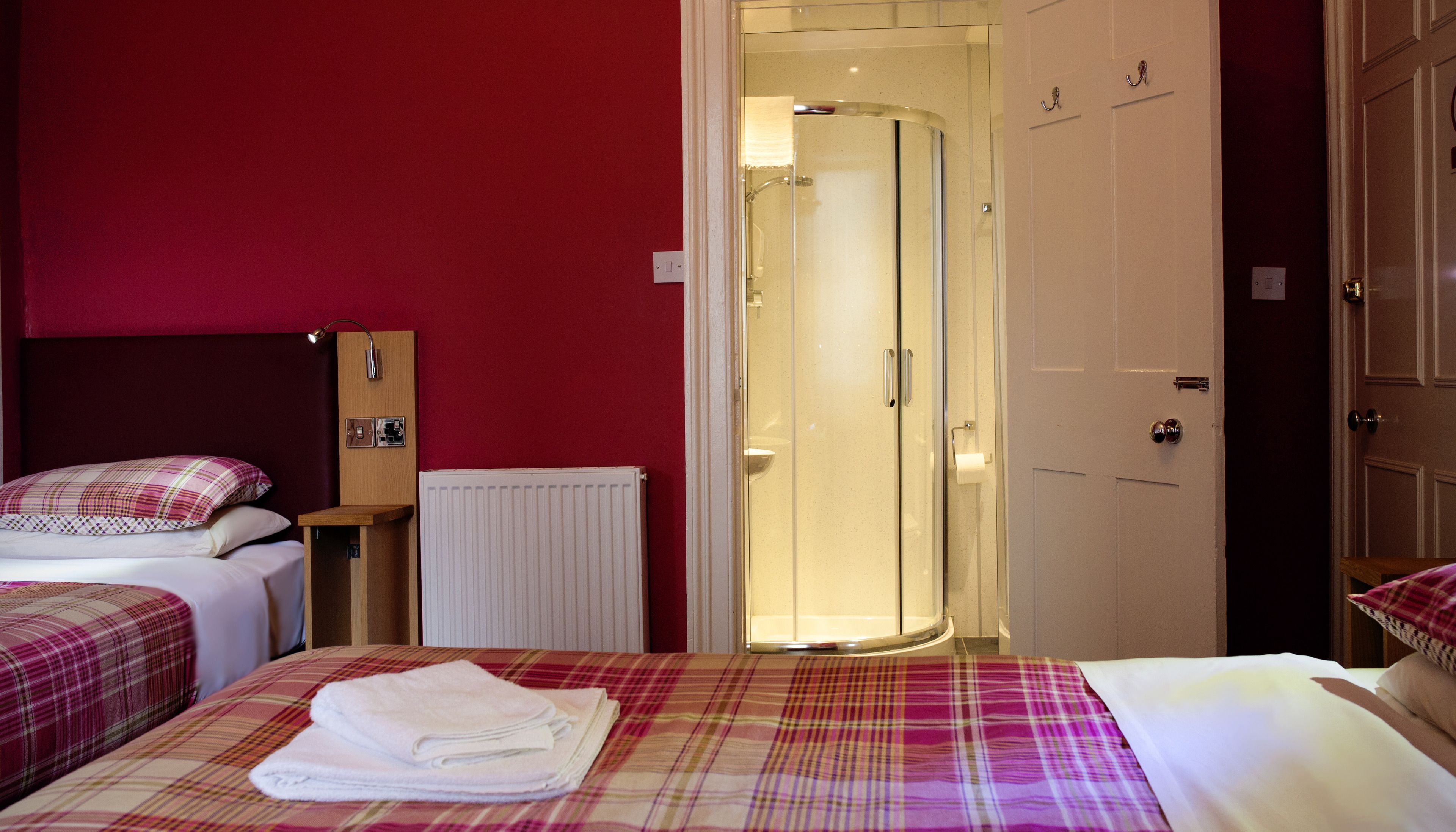 Une chambre d’hôtel avec deux lits, un mur rouge, une porte de douche en verre et un radiateur blanc.