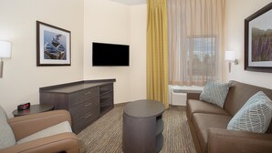 A hotel room with a flat-screen TV, a sofa, a coffee table, a TV stand, and a window with curtains. - Candlewood Suites Cheyenne by IHG (Cheyenne)