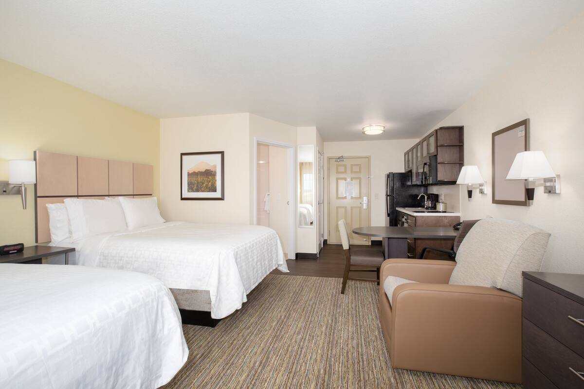 A hotel room with two beds, a desk, a chair, a kitchenette, and a bathroom.