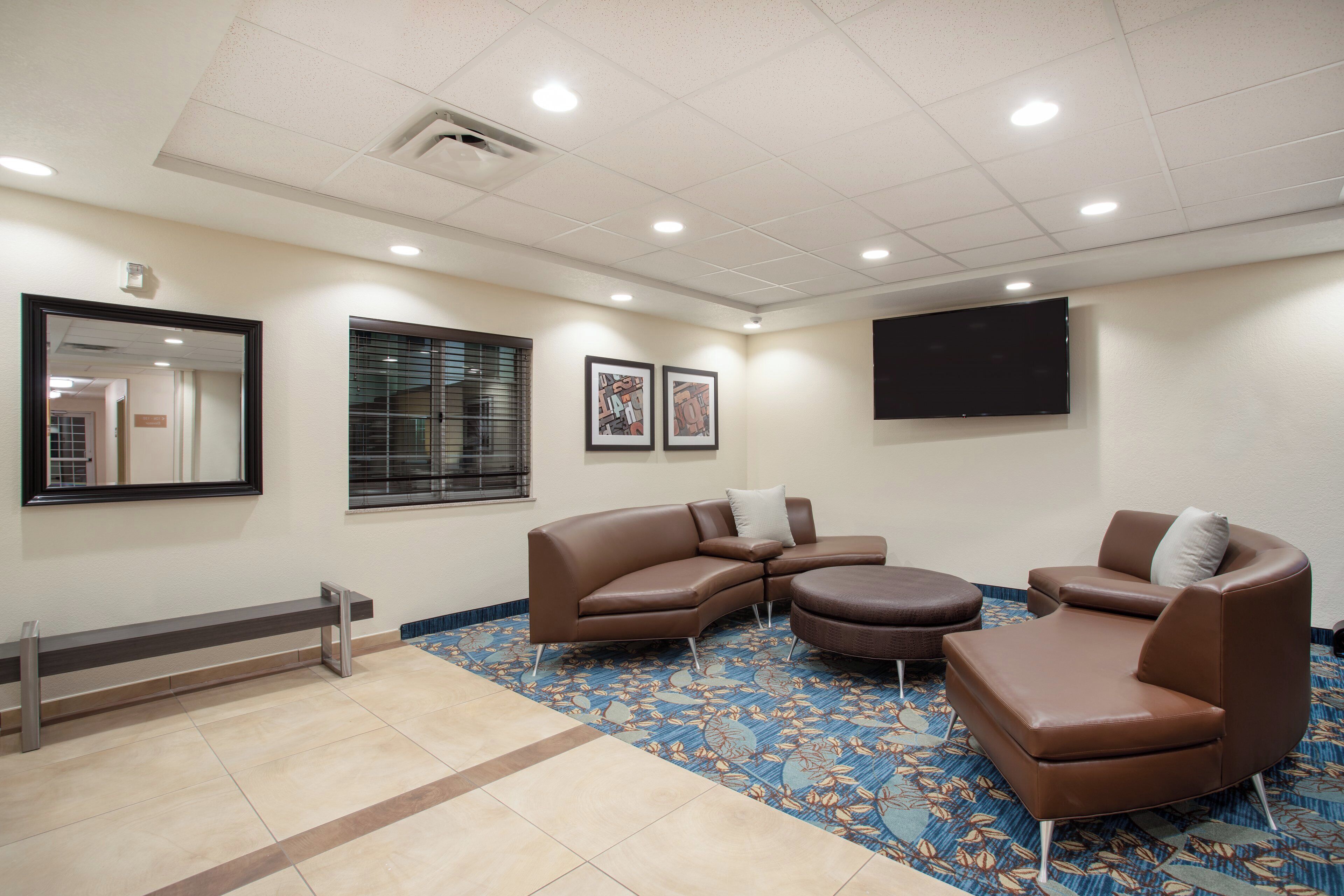 A hotel room with a sectional sofa, a small round ottoman, a bench, a flat-screen TV, and artwork on the walls.
