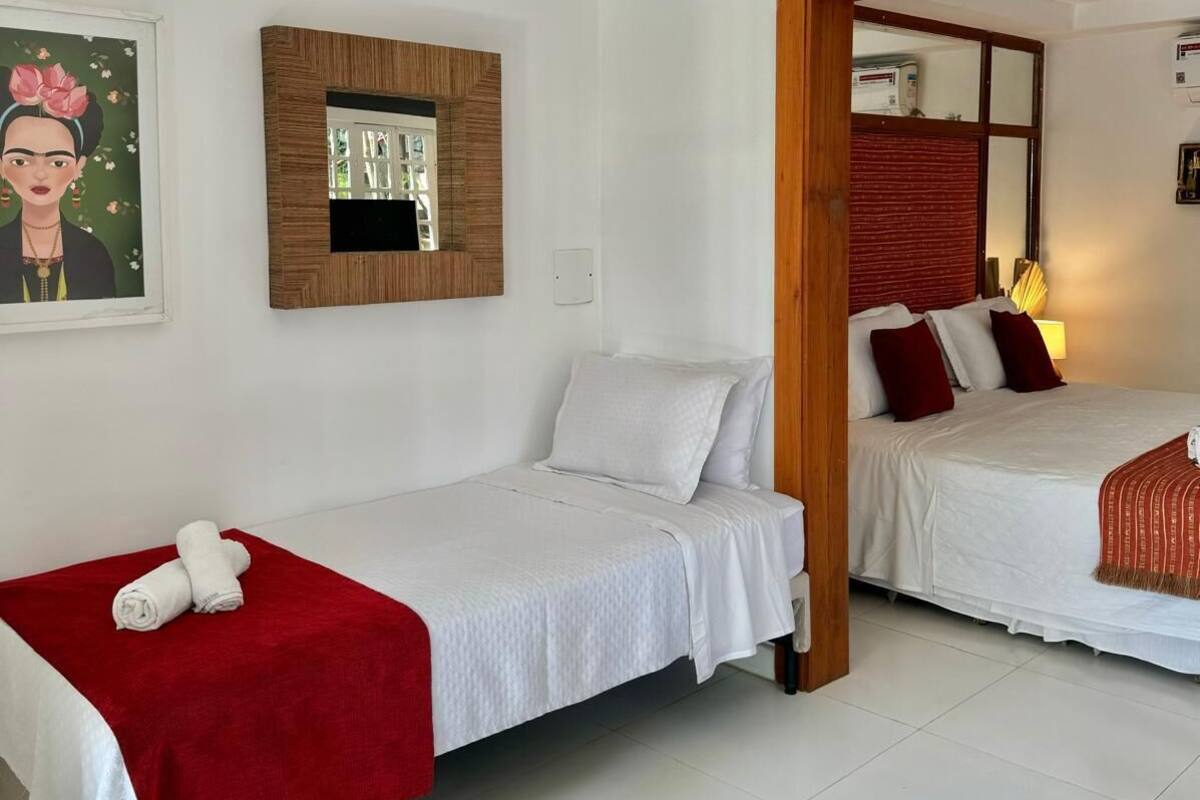 Habitación de hotel con dos camas, una con una colcha roja y la otra con almohadas rojas y blancas. Tiene un cabecero de madera, un cuadro en la pared y una mesita con una lámpara.