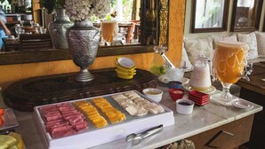 Una mesa de buffet con diversos alimentos y bebidas, incluyendo una bandeja de frutas, una selección de carnes y una variedad de refrescos en diferentes vasos.