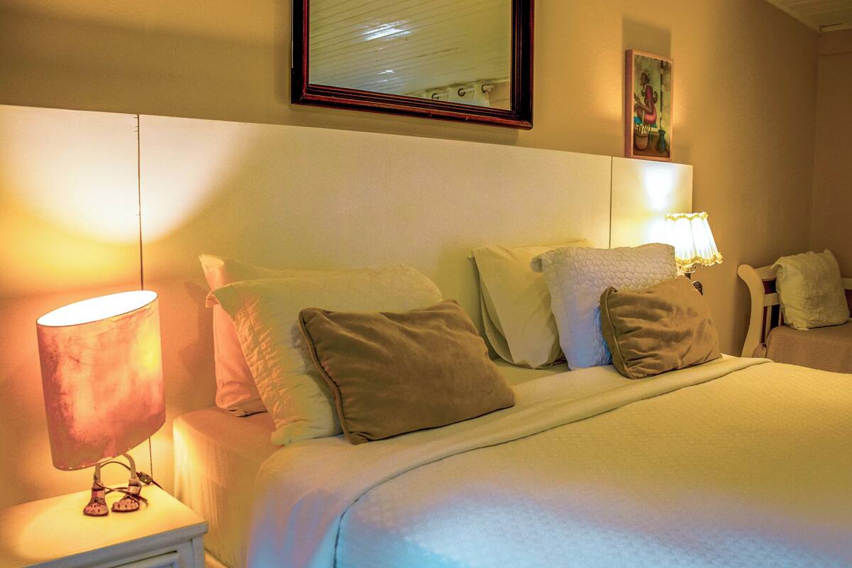 Habitación de hotel con una cama bien hecha, una lámpara de noche, una mesita de noche con un cajón y un cuadro en la pared.