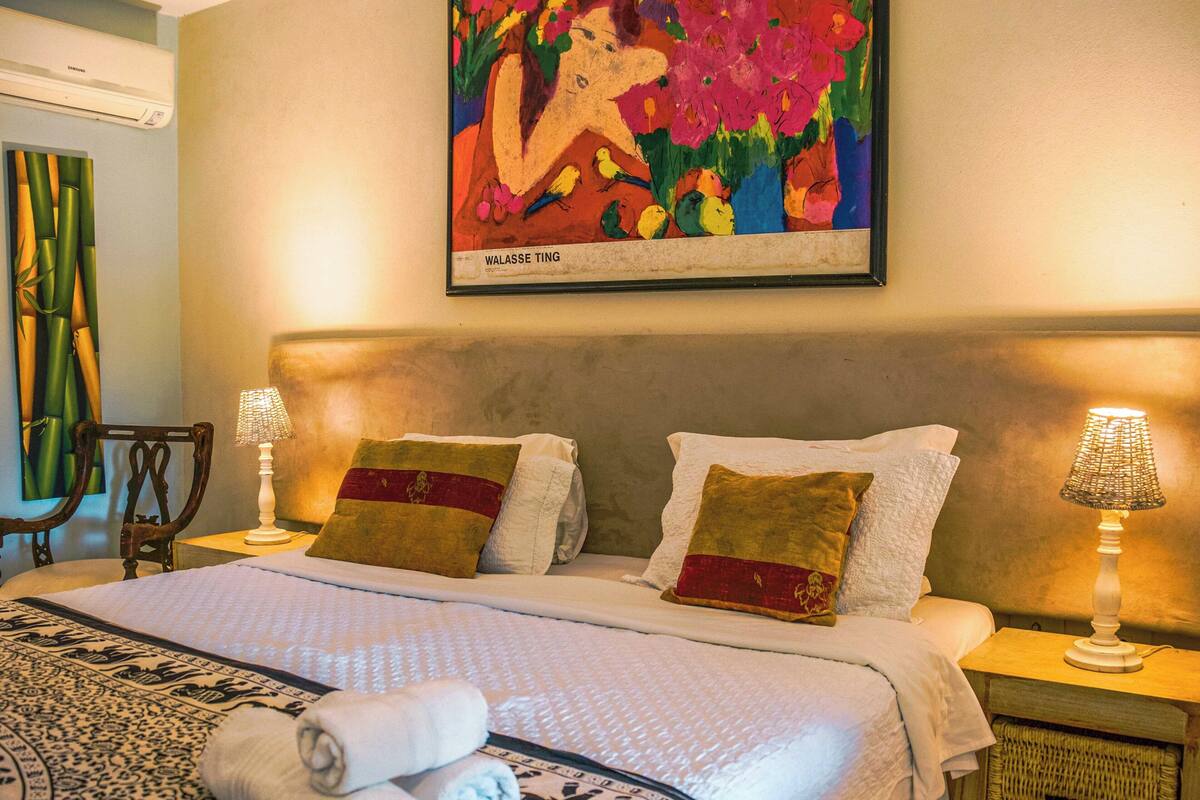 Una habitación de hotel con una cama, dos almohadas, una mesita de noche con una lámpara, una silla, un aire acondicionado instalado en la pared y un cuadro enmarcado.