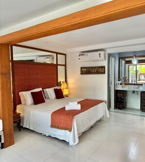 Une chambre d’hôtel avec un grand lit, une tête de lit en bois, une table de chevet, une lampe, un miroir, une fenêtre et un coin cuisine.
