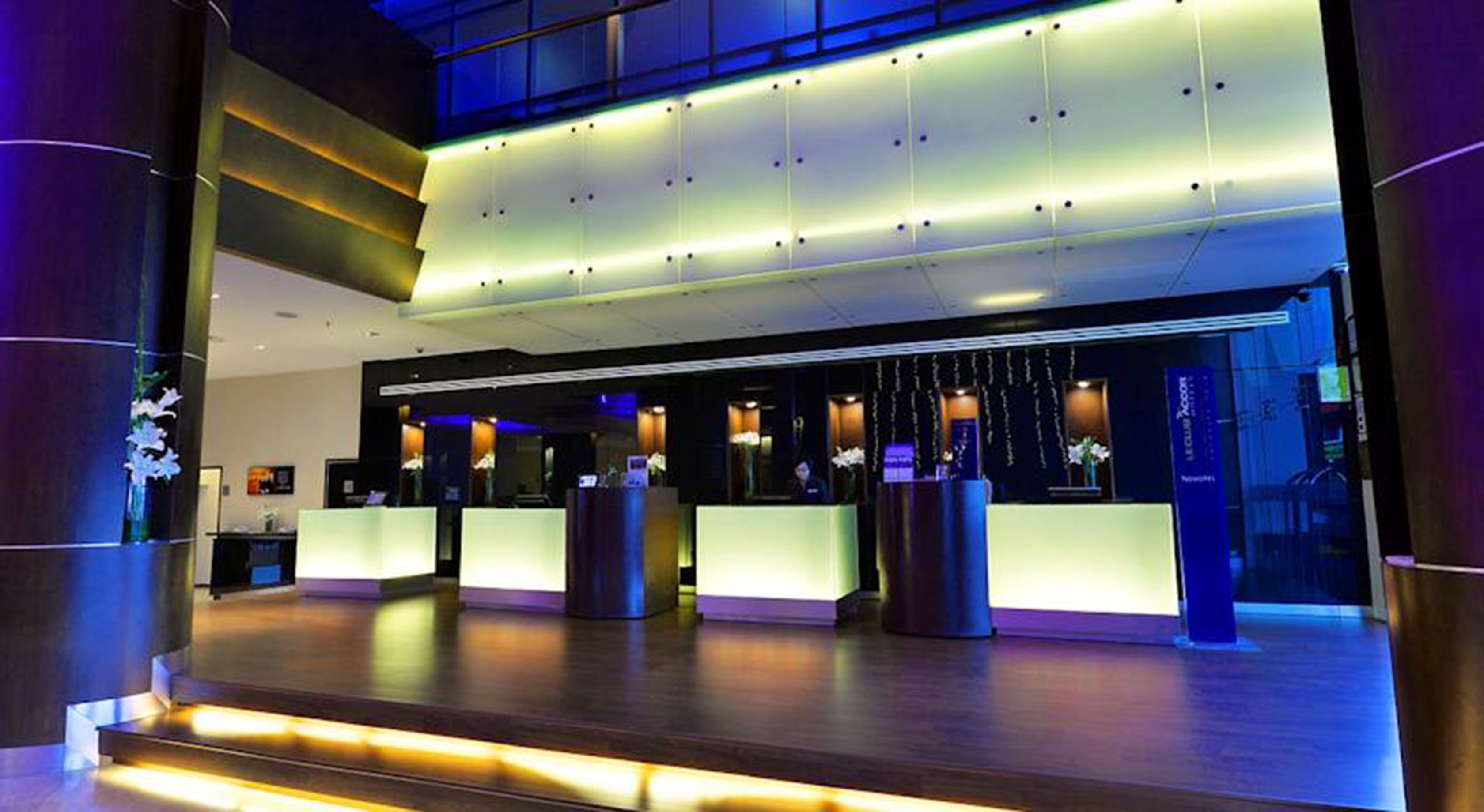 A modern hotel lobby with a reception desk, a large seating area, and a staircase leading to an upper level.