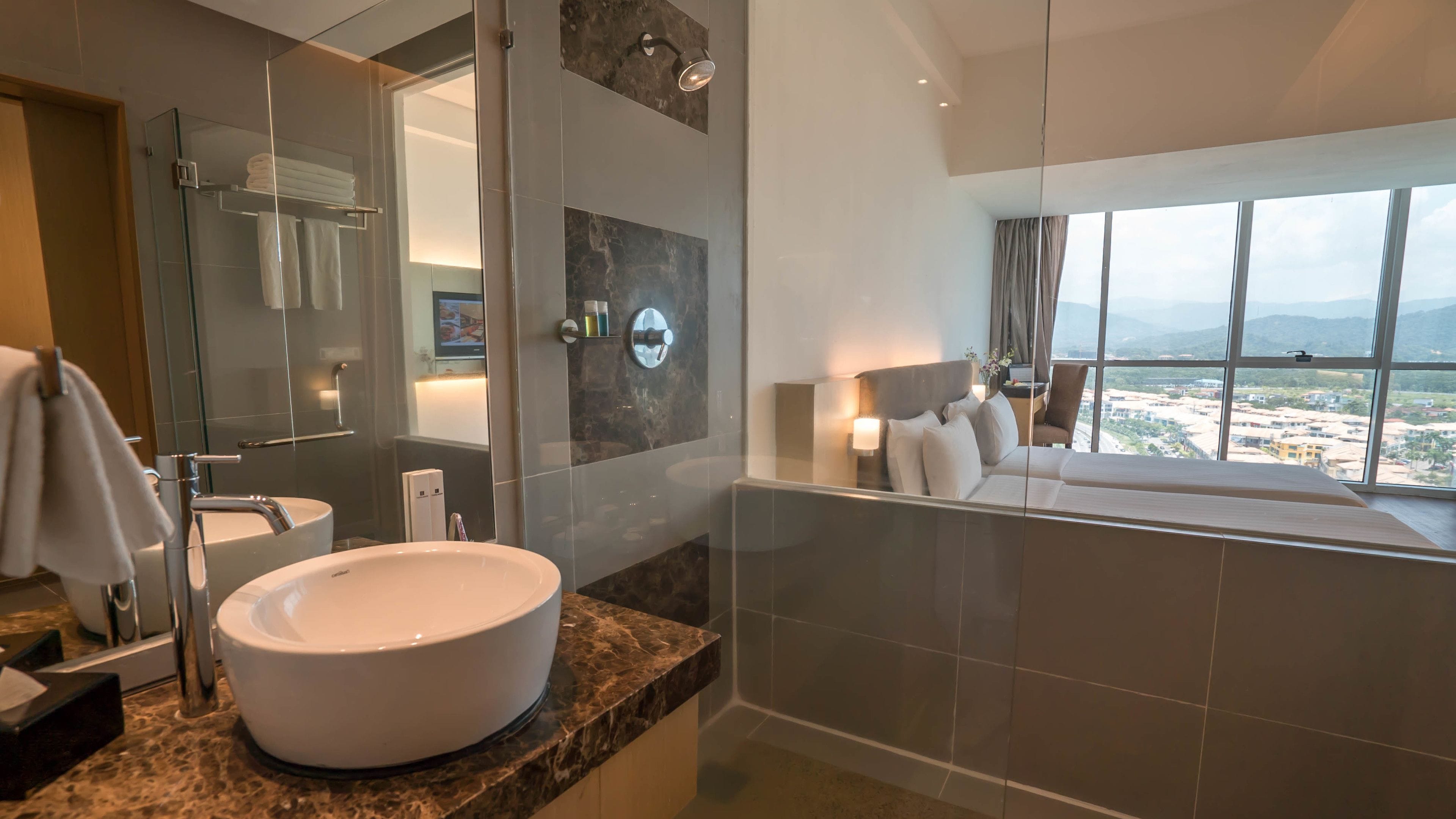 A modern bathroom with a large white sink, a glass-enclosed shower, and a view of a cityscape through a large window.