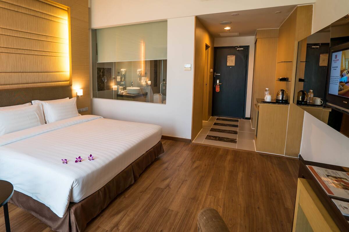 A hotel room with a large bed, a small table with a glass of water, a TV, and a kitchenette area.