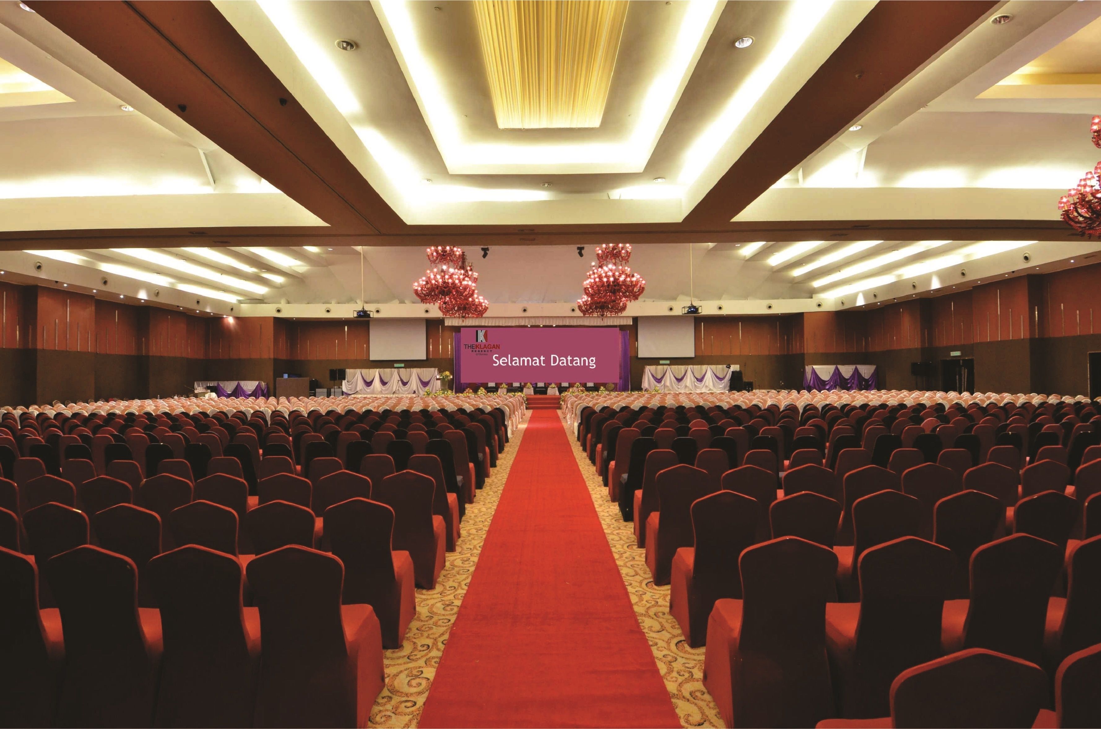 Un amplio salón de conferencias con filas de sillas, una alfombra roja y un escenario con un estandarte morado.