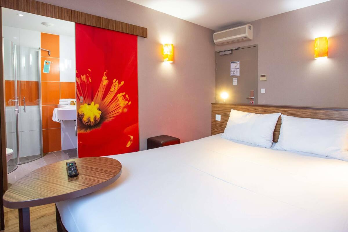 A hotel room with a bed, a round table with a remote control, a bathroom with a shower, and two wall-mounted lights.