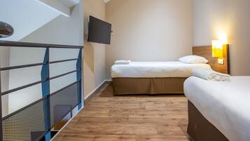 Una habitación de hotel con piso de madera, una cama con ropa blanca, un televisor montado en la pared y una mesita de noche con lámpara.