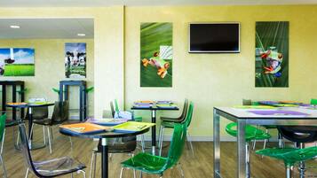 A brightly lit indoor space with multiple tables and chairs, artwork on the walls, and a flat-screen TV mounted above a table.