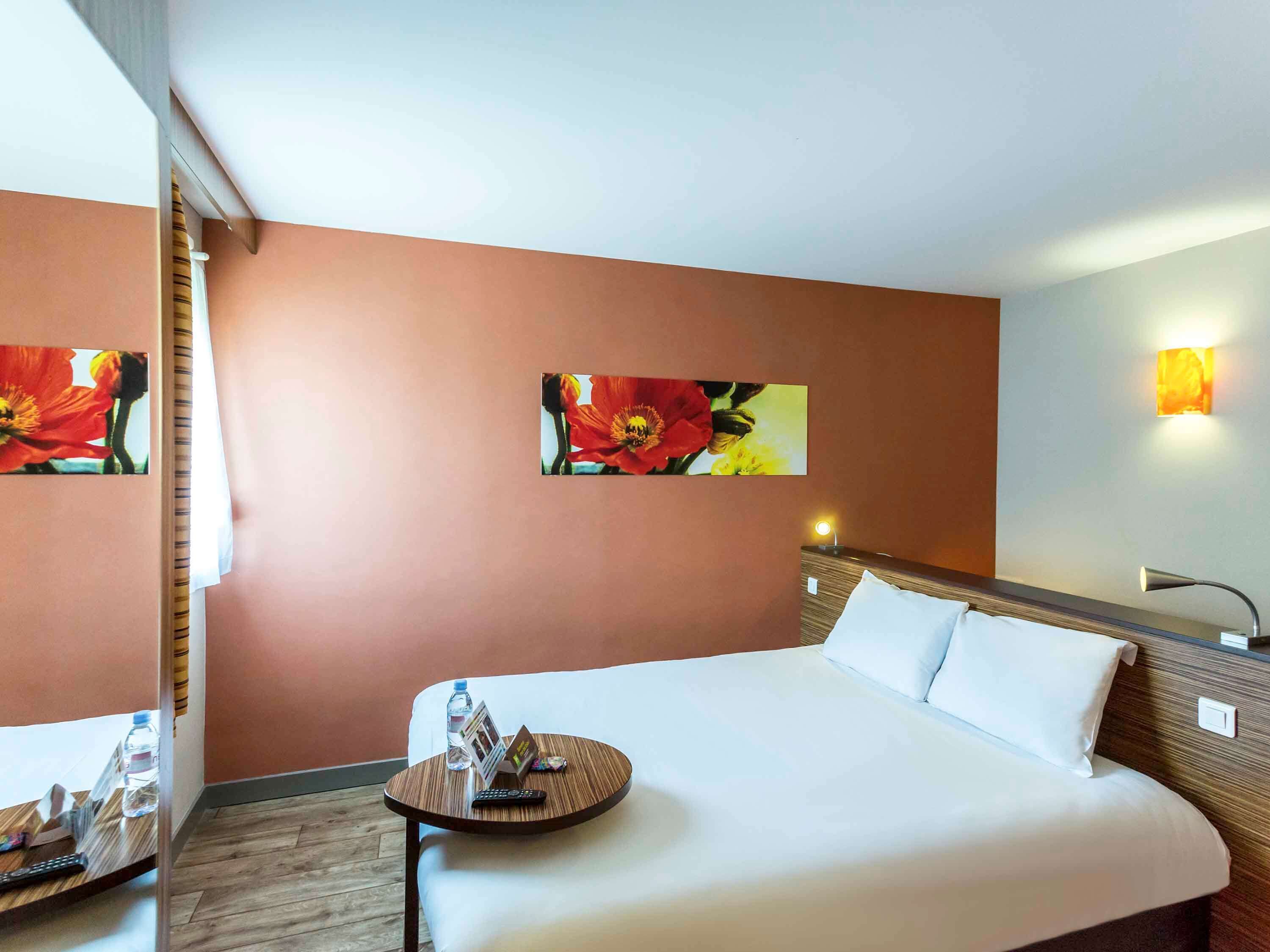 Ein Hotelzimmer mit einem großen Bett, einem Nachttisch mit einer Wasserflasche, einer Fernbedienung und einer Zeitschrift. Es befinden sich zwei Wandlampen und ein Blumenbild an der Wand.