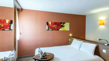 Ein Hotelzimmer mit einem großen Bett, einem Nachttisch mit einer Wasserflasche, einer Fernbedienung und einer Zeitschrift. Es befinden sich zwei Wandlampen und ein Blumenbild an der Wand.