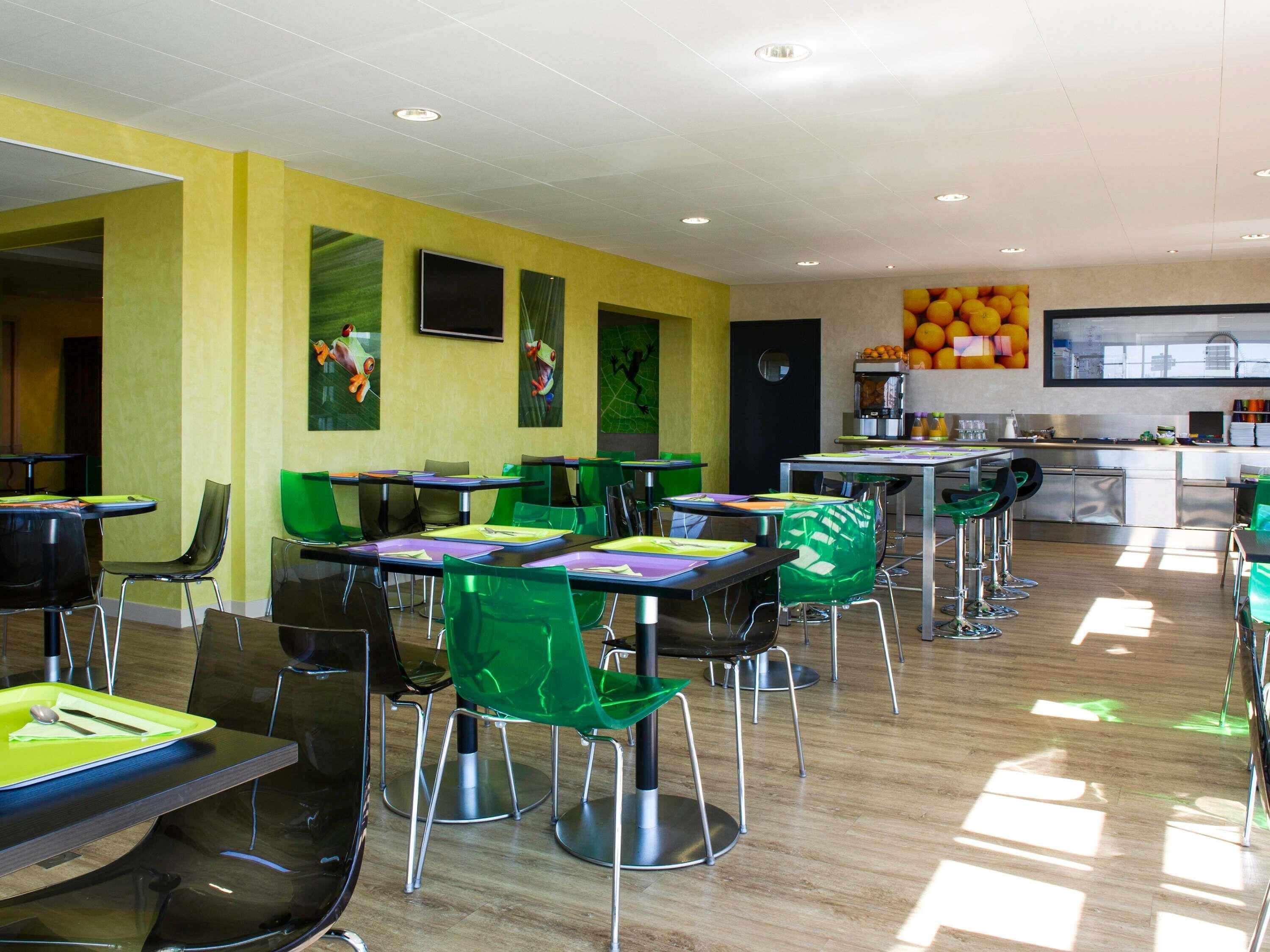 Un comedor moderno con sillas y mesas verdes, una zona de cocina con una exposición de frutas, y una pared de color amarillo brillante.