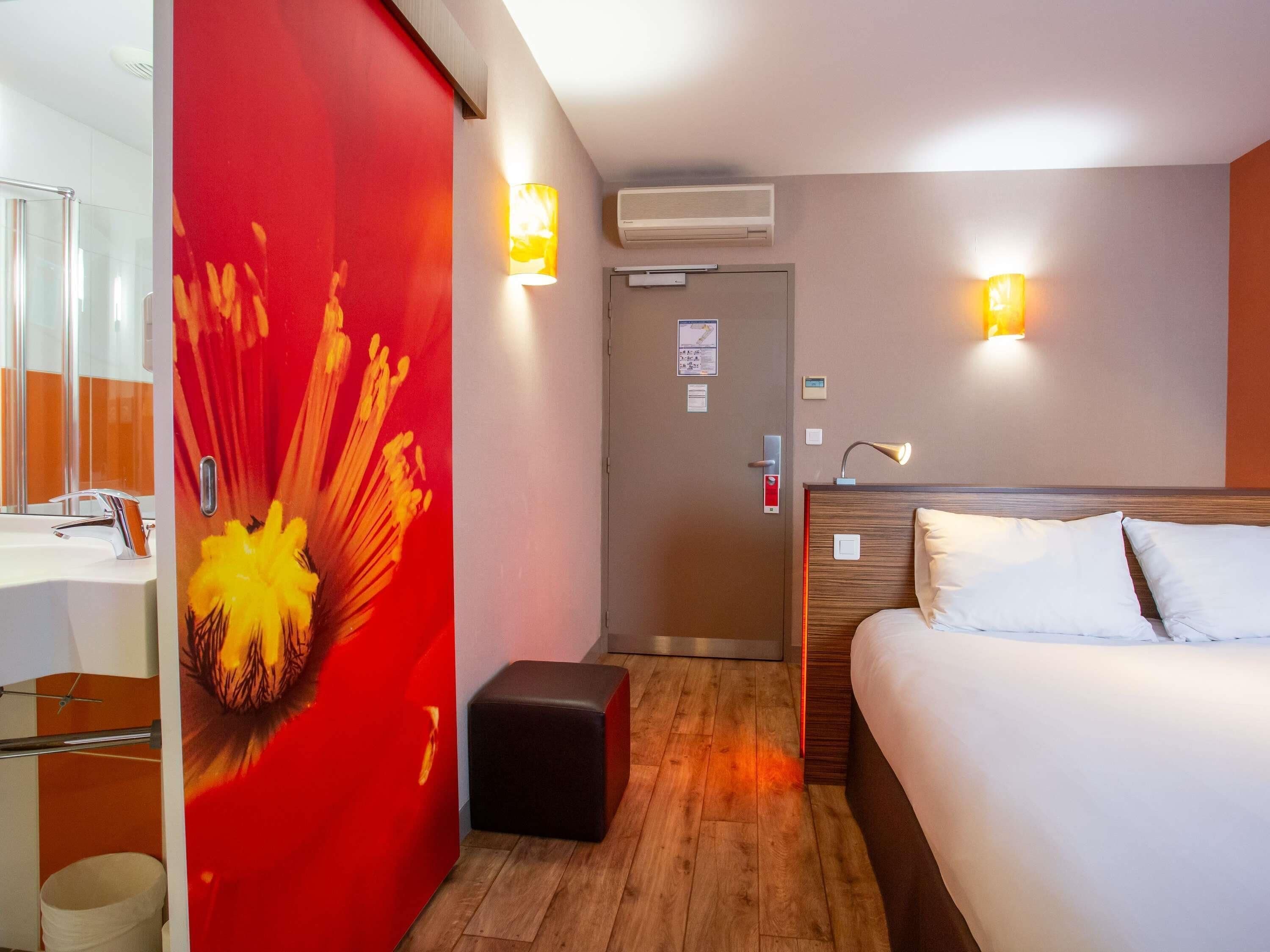 A hotel room with a bed, a bedside table, a mirror, a red accent wall, and two wall-mounted lights.