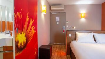 Habitación de hotel con cama, mesita de noche, espejo, pared de acento roja y dos luces fijadas a la pared.