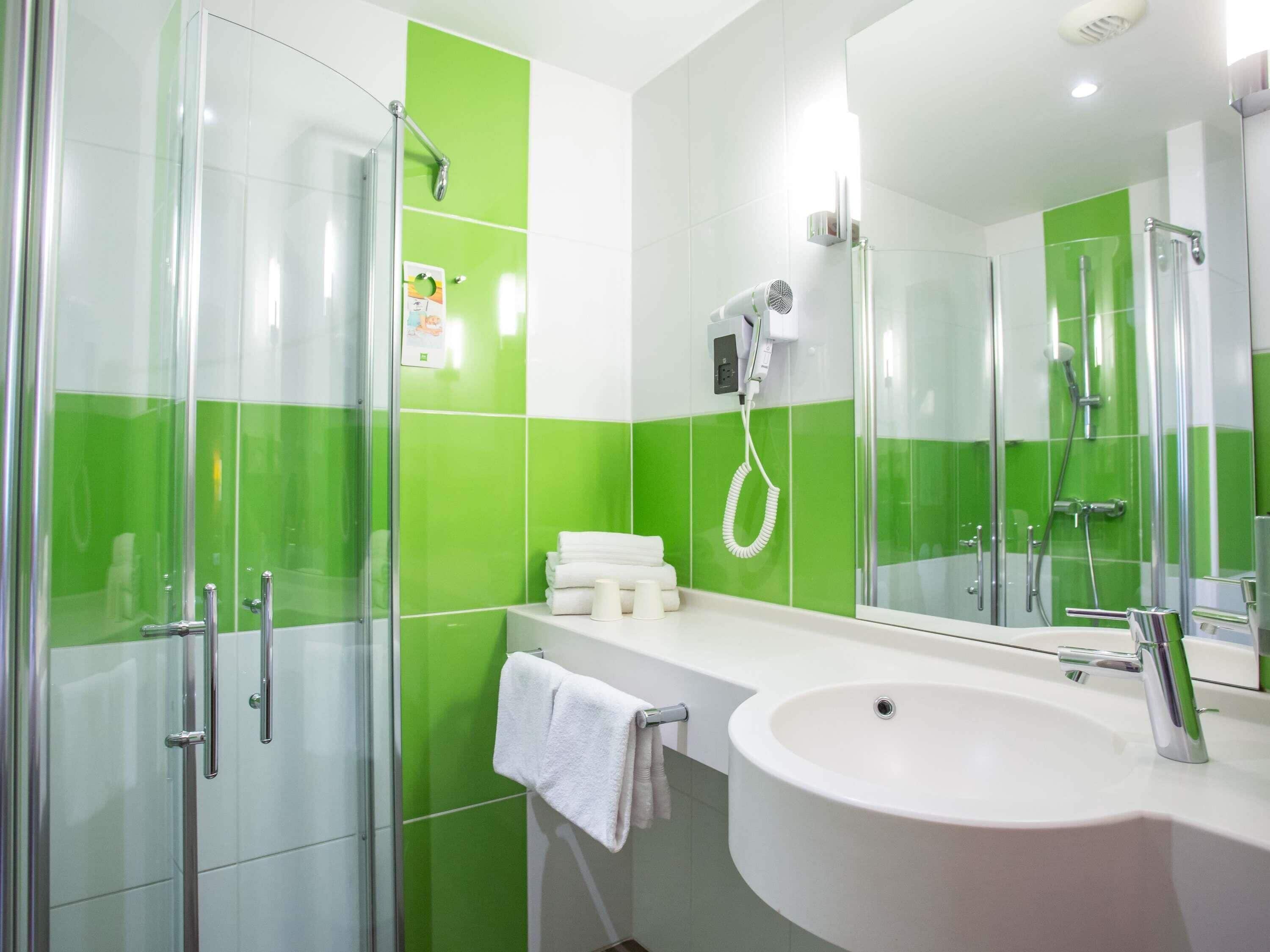 Ein modernes Badezimmer mit Duschkabine aus Glas, einem weißen Waschbecken und einer grün gefliesten Wand, sowie einem Haartrockner an der Wand.