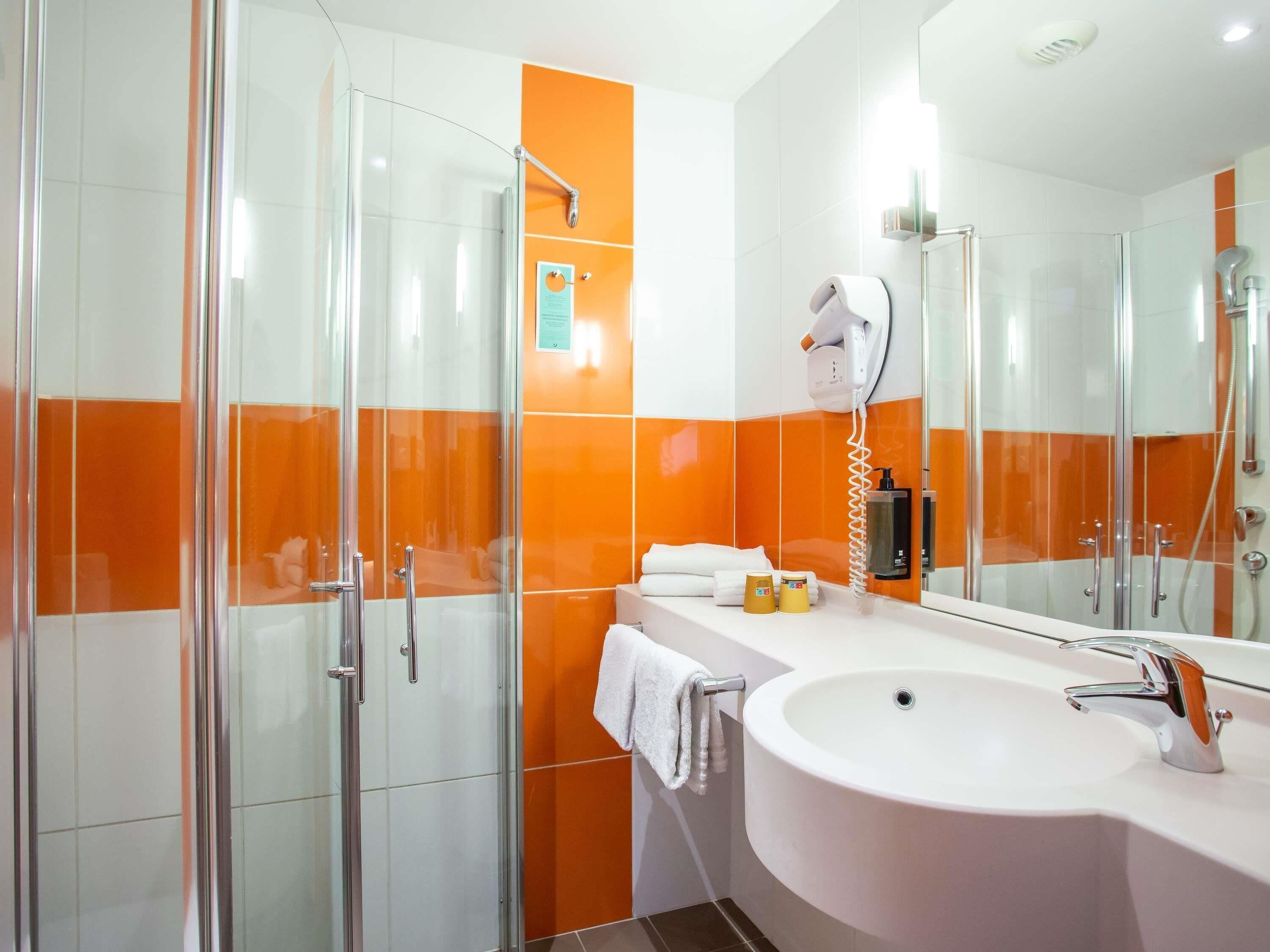 Un baño moderno con cabina de ducha de vidrio, un lavamanos blanco con grifo cromado y una pared con azulejos naranja.