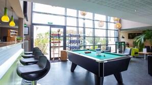 A pool table in a well-lit indoor space with large windows, bar area, and seating. - ibis Styles Bourges (Bourges)