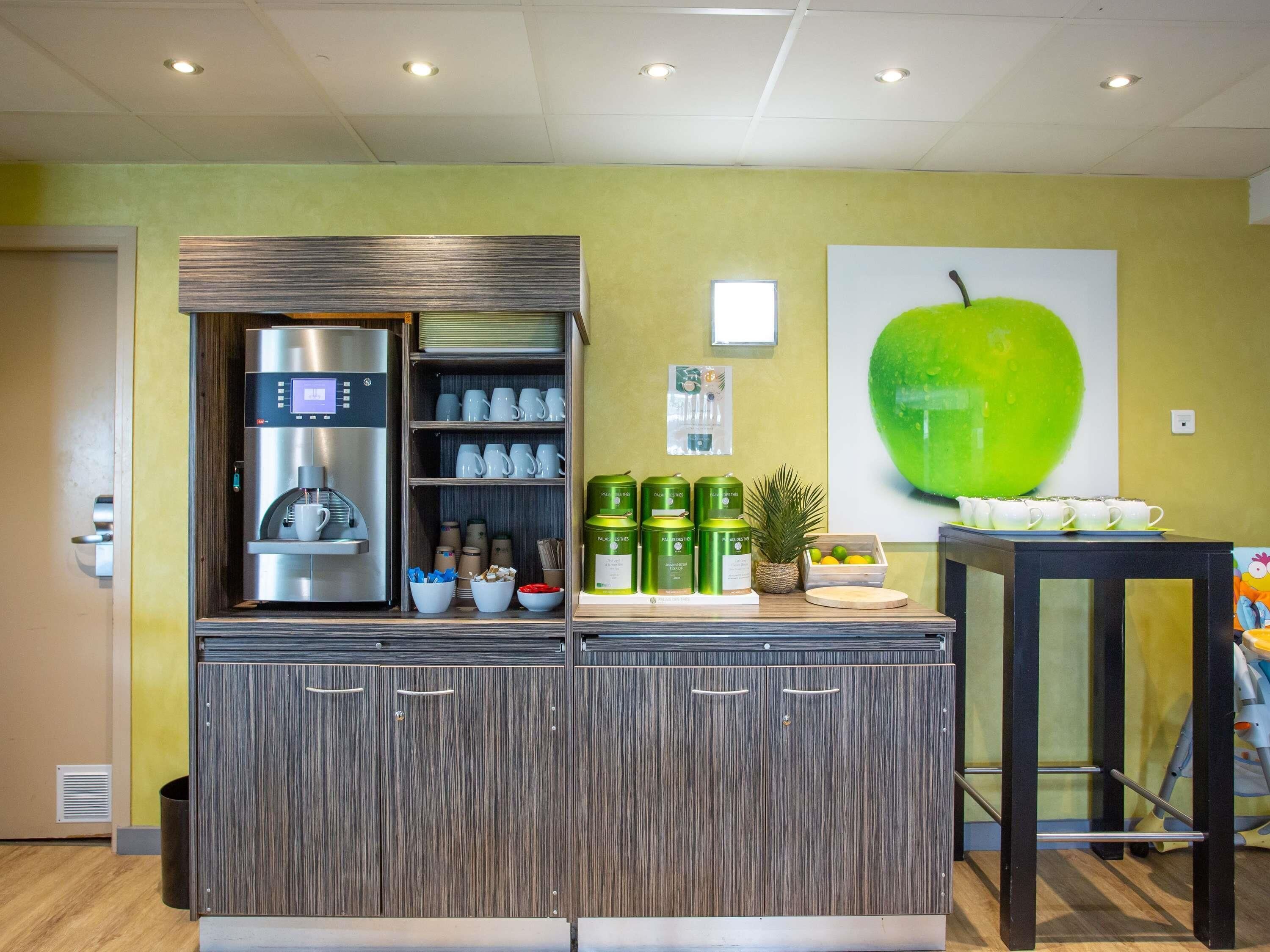 Una kitchenette con una cafetera, estantes con tazas y un cuadro grande de una manzana verde en la pared.