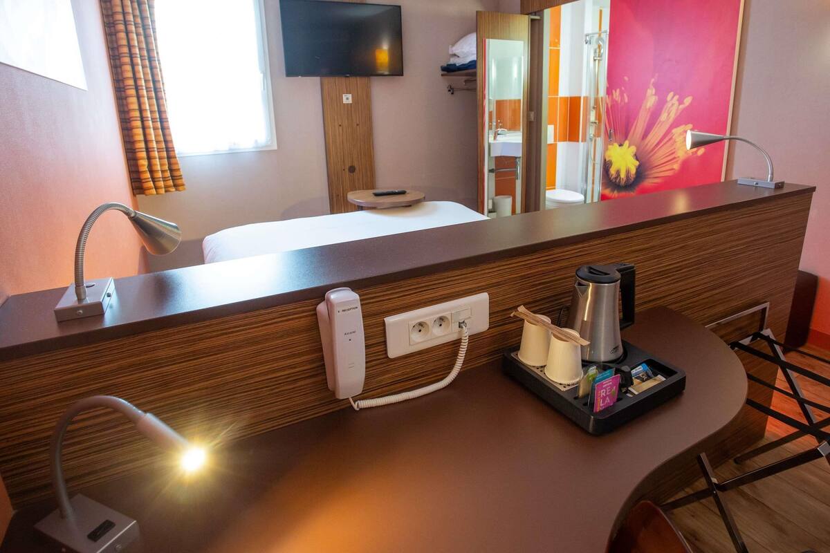 Ett hotellrum med en receptionsdisk i trä, en platt-TV som hänger ovanför, en spegel, ett sängbord med en lampa och en kaffebryggare.