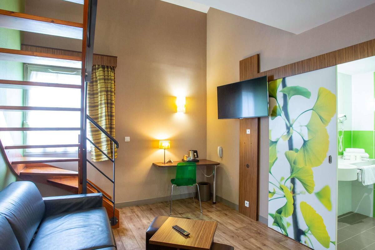 Ett hotellrum med en soffa, ett litet bord med en fjärrkontroll, ett skrivbord med en lampa, en tv som är monterad på väggen och ett badrum med en grön vägg och en spegel.