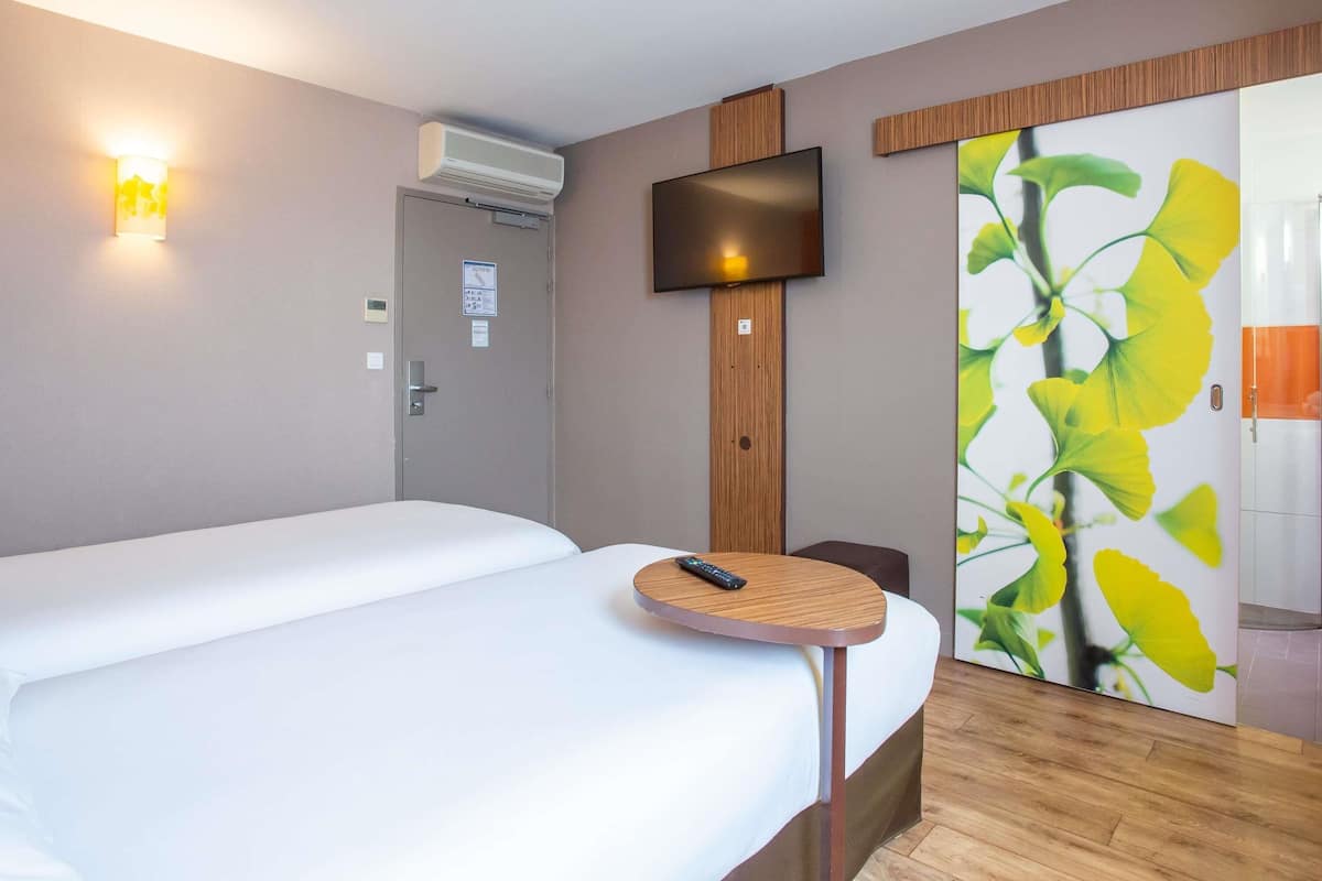 A hotel room with a bed, a TV mounted on the wall, a small table with a remote control, and a door with a plant wallpaper.