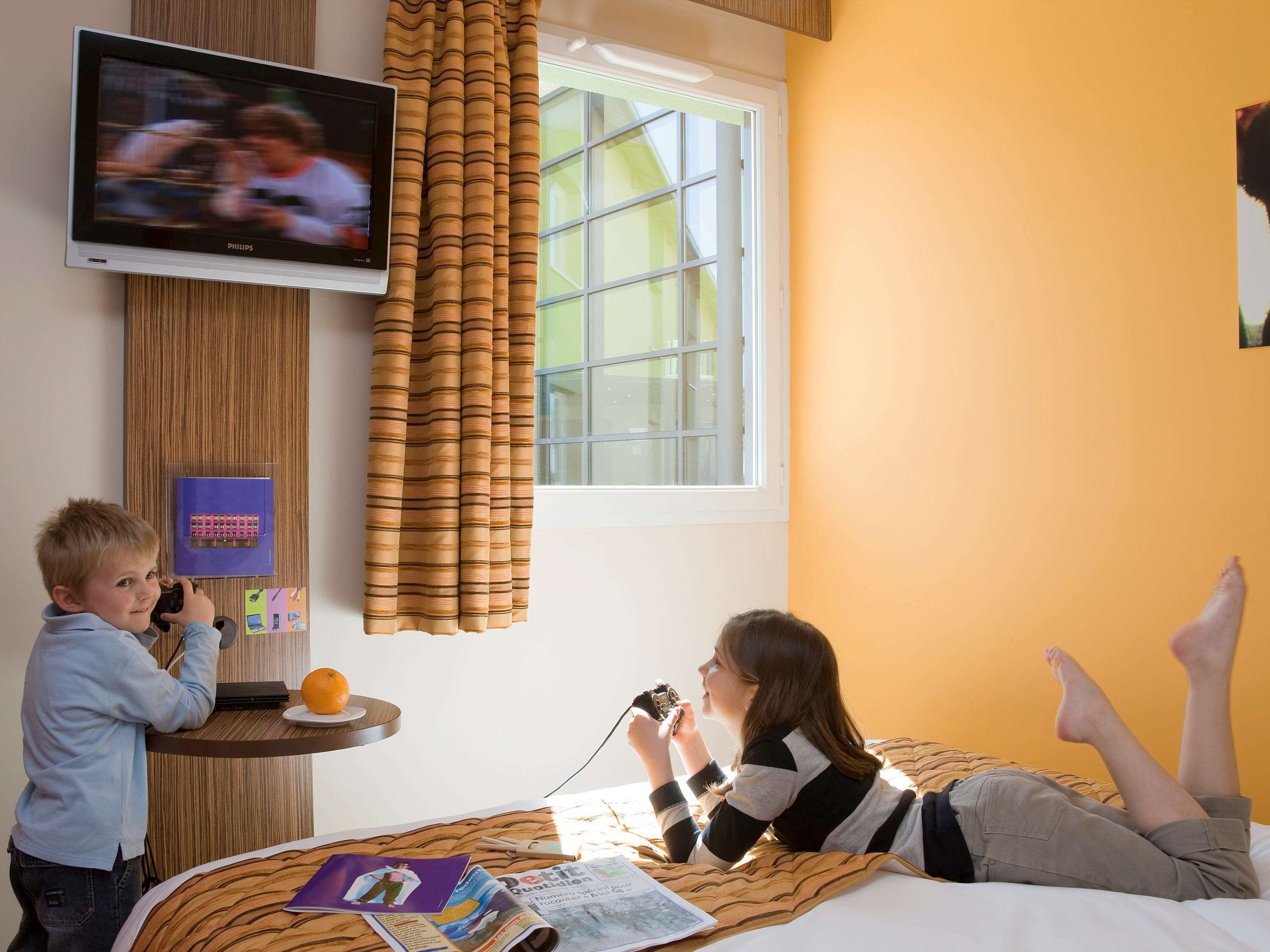 A hotel room with a bed, a television mounted on the wall, a window with curtains, and a small table with a magazine and an orange.