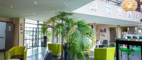 Un espacio interior moderno con áreas de estar, una planta grande en maceta y una zona de bar con mostrador y luces colgantes.
