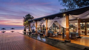 A waterfront restaurant with outdoor seating, a pool, and a view of the ocean at sunset.