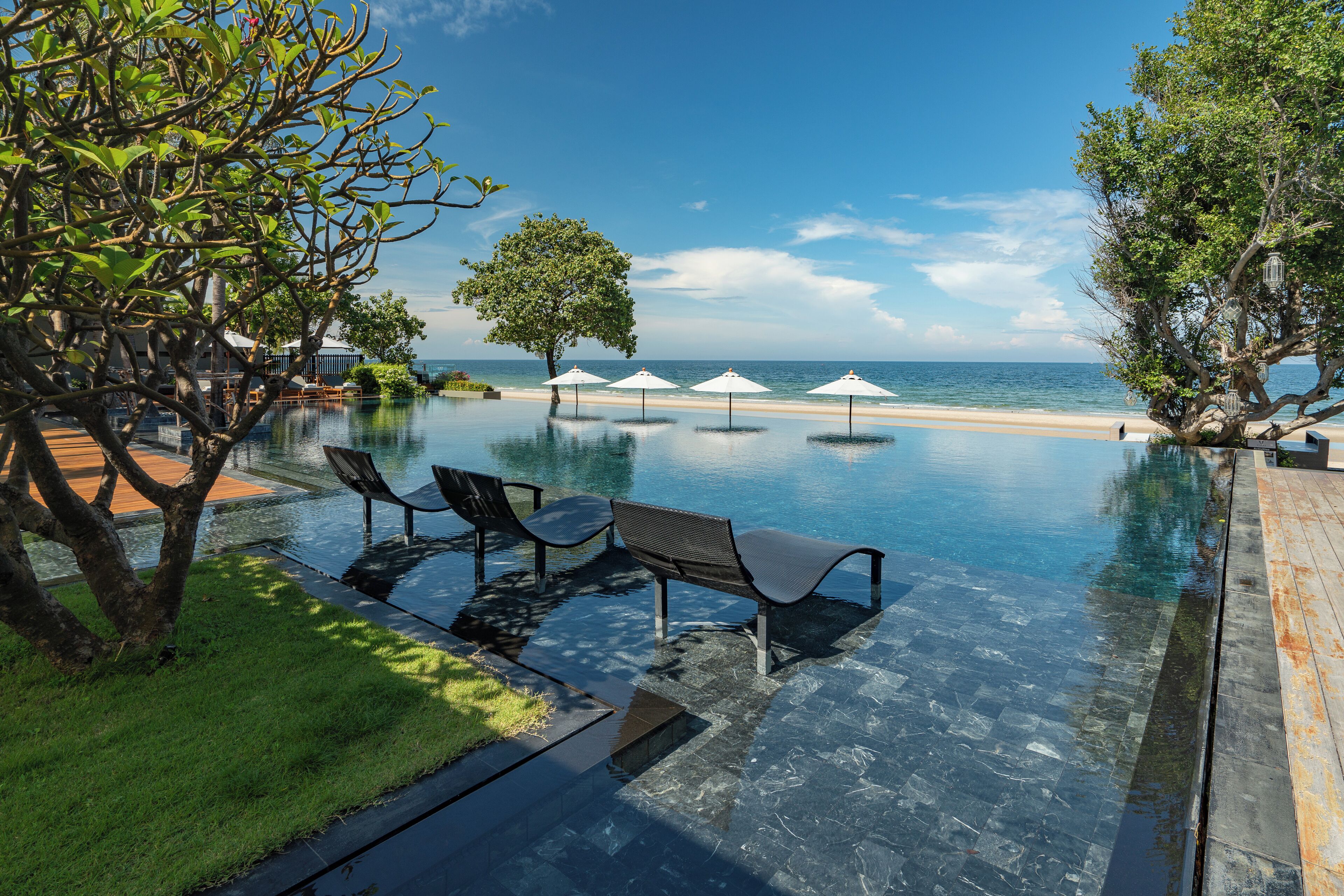 An infinity pool with clear water, surrounded by lounge chairs and umbrellas, overlooking a beach.